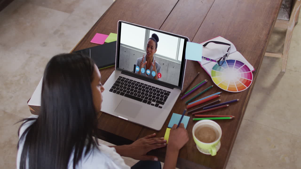 mujer caucásica usando una computadora portátil en videollamada con una colega y tomando notas