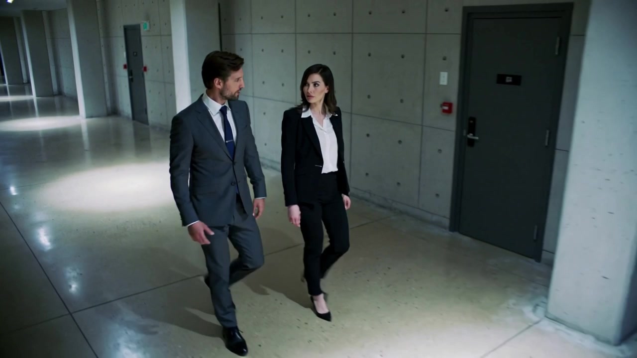 Due colleghi camminano e parlano in un corridoio di cemento, indossando un abito e un blazer.