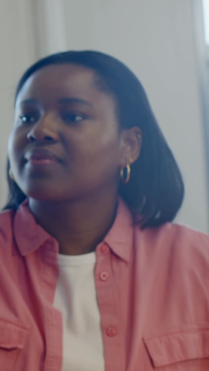 Vertical video: Listening woman in pink top gesturing as offscreen voice starting in room by window