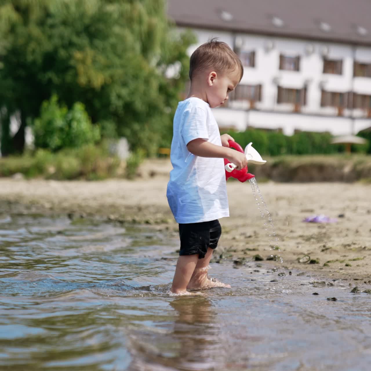 Little kid stands in river pouring water from his red watering can. Cute Caucasian toddler playing on the beach in summer