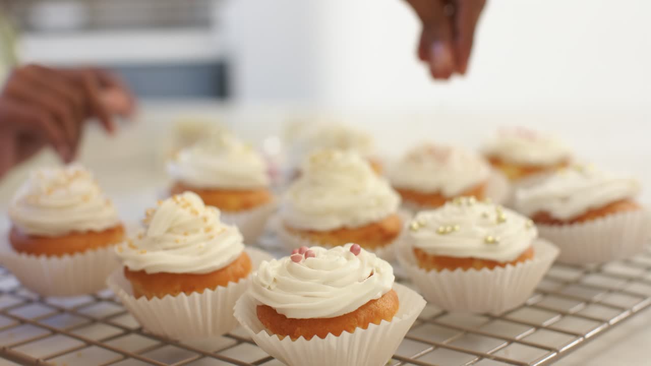 Mãos entrando pela esquerda estão espalhando confeitos dourados e pérolas rosas em cupcakes com cobertura para decorar