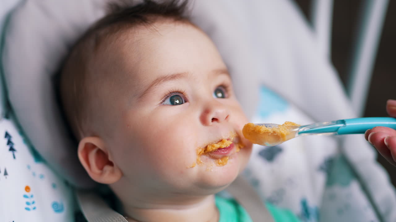 Nice kid eating porridge from spoon. Adorable toddler sticks out tongue and waves his hands. Close up.
