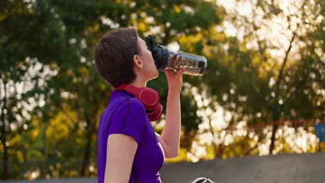 side view of a happy girl in a purple top and red headphones with a short haircut drinks water and specially bottles in a skate park in summer