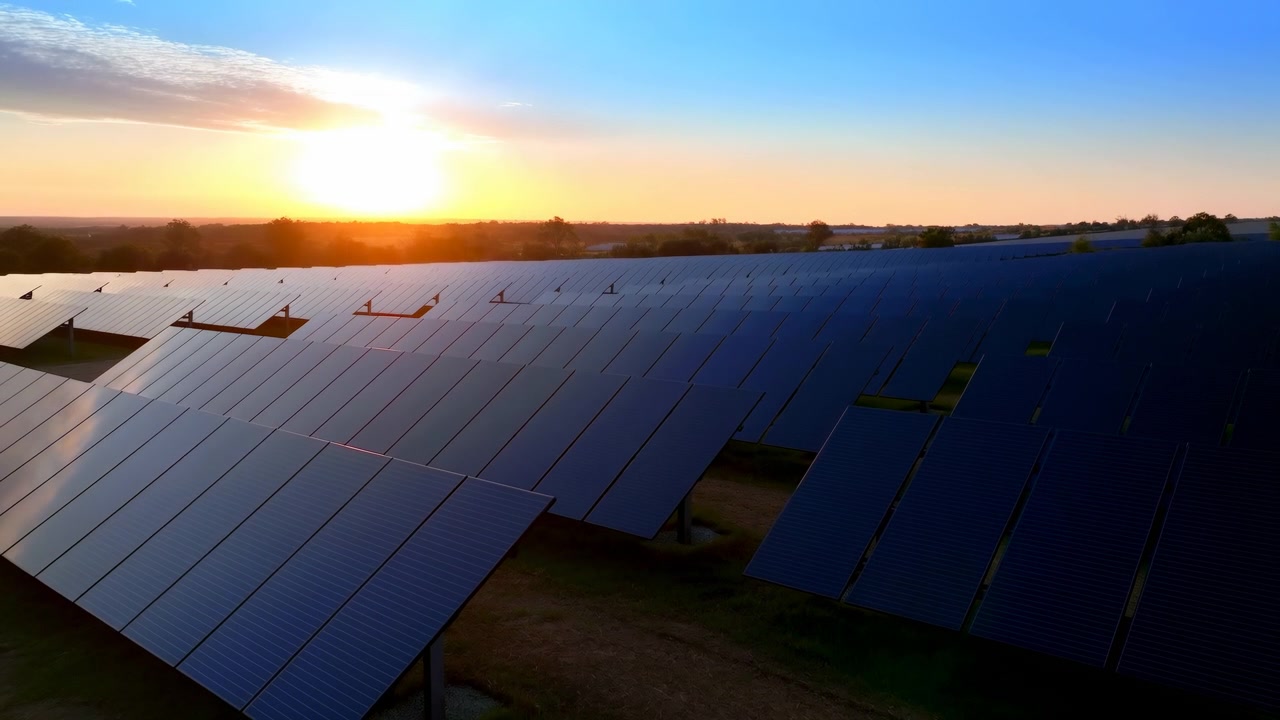 Ampia scena panoramica di un grande impianto fotovoltaico a terra al tramonto con più file di pannelli solari riflettenti blu scuro angolati su supporti metallici che si estendono verso l'orizzonte lontano in un'area rurale