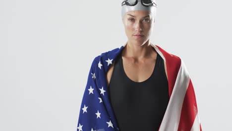 joven atleta femenina caucásica nadadora envuelta en una bandera estadounidense se encuentra con confianza