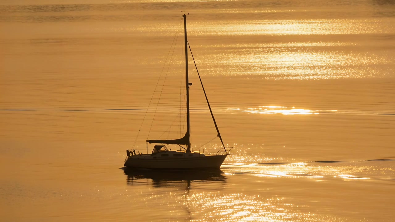Barca a vela che veleggia con vela ammainata, alla deriva in una leggera brezza su una baia dorata all'alba, lasciando una scia.