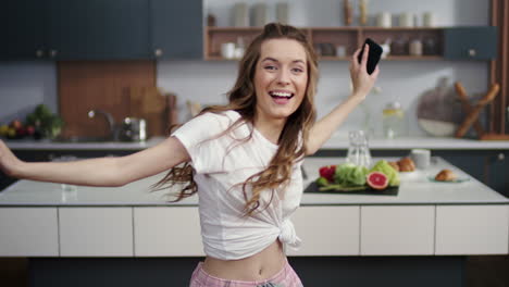 Woman-dancing-with-mobile-phone-in-hand-at-home.-Cute-girl-smiling-to-camera