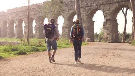 young couple backpackers tourists walking near roman aqueduct arches in parco degli acquedotti park ruins in rome at sunrise with guitar and sleeping bag slow motion steadycam