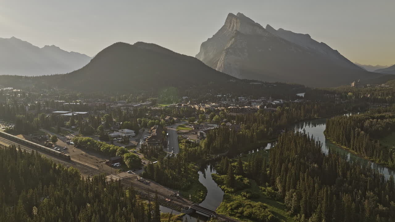 banff ab canada aerial v33 cinematic flyover capturing scenic landscape of train crossing quaint town by the bow river and rundle mountain ranges at sunrise - снят с помощью mavic 3 pro cine - июль 2023 года