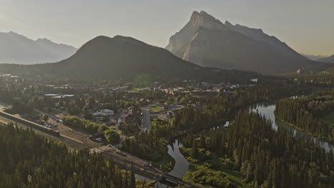 banff ab canada aerial v33 cinematic flyover capturing scenic landscape of train crossing quaint town by the bow river and rundle mountain ranges at sunrise - shot with mavic 3 pro cine - july 2023