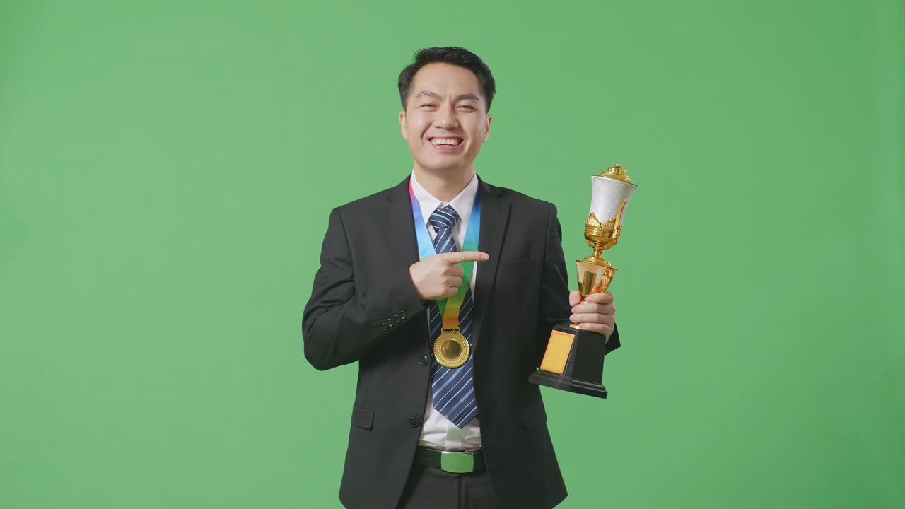 슈트와 타이를 입은 아시아 사업가, 손에 황금 트로피를 가리키며, 스튜디오의 녹색 스크린 배경에서 첫 번째 우승자로 자랑스럽게 승리합니다.