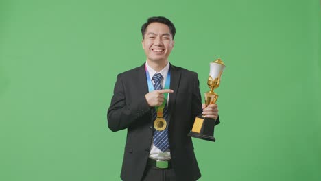 슈트와 타이를 입은 아시아 사업가, 손에 황금 트로피를 가리키며, 스튜디오의 녹색 스크린 배경에서 첫 번째 우승자로 자랑스럽게 승리합니다.