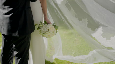 Close-up-of-a-bride-holding-a-bouquet-while-standing-with-the-groom,-her-veil-billowing-in-the-wind