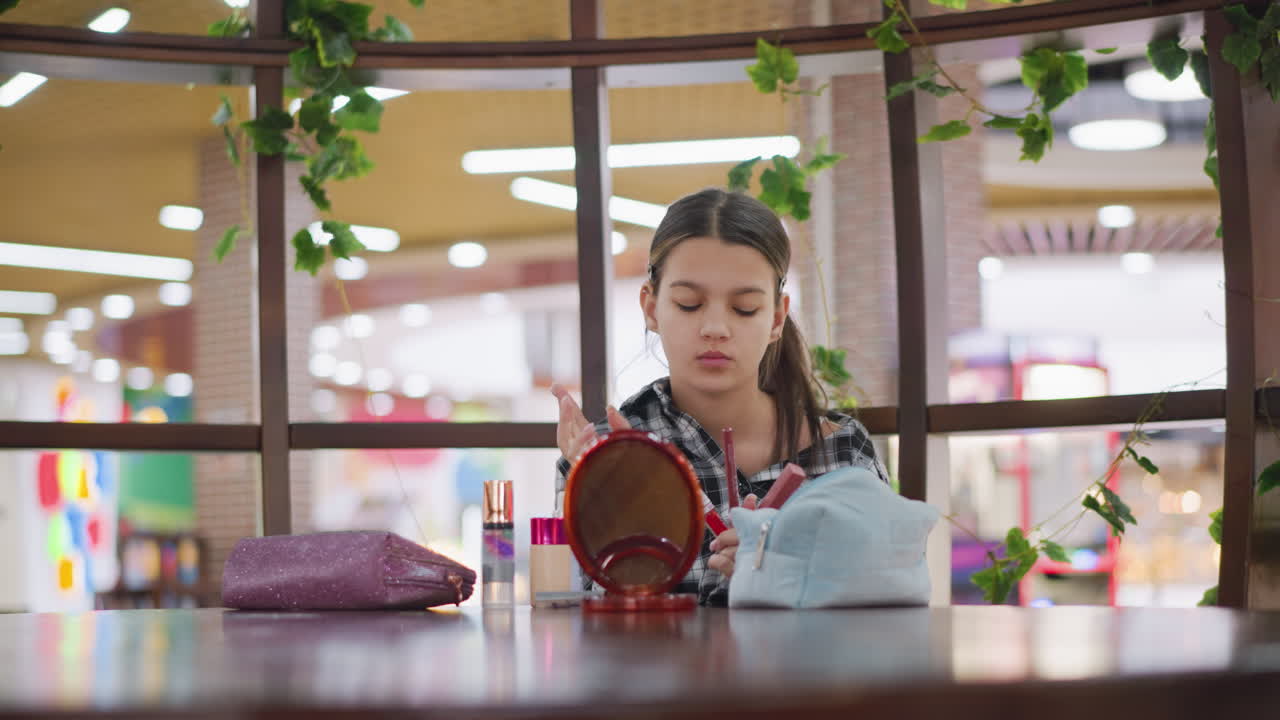 젊은 여자가 거울 앞에 앉아 손에 있는 화장품을 조심스럽게 식별하고, 화장품과 세련된 장식품으로 둘러싸여 적절한 미용품을 선택하는 데 집중합니다.