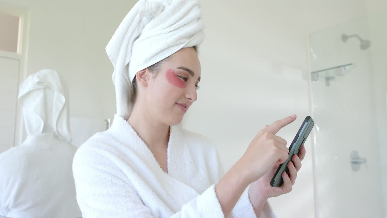 Donna caucasica felice con patch per gli occhi che usa lo smartphone in bagno al rallentatore