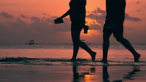 couple walks barefoot in shallow sea water at bali sunset - outrigger old boat in silhouette anchored floating in ocean in background