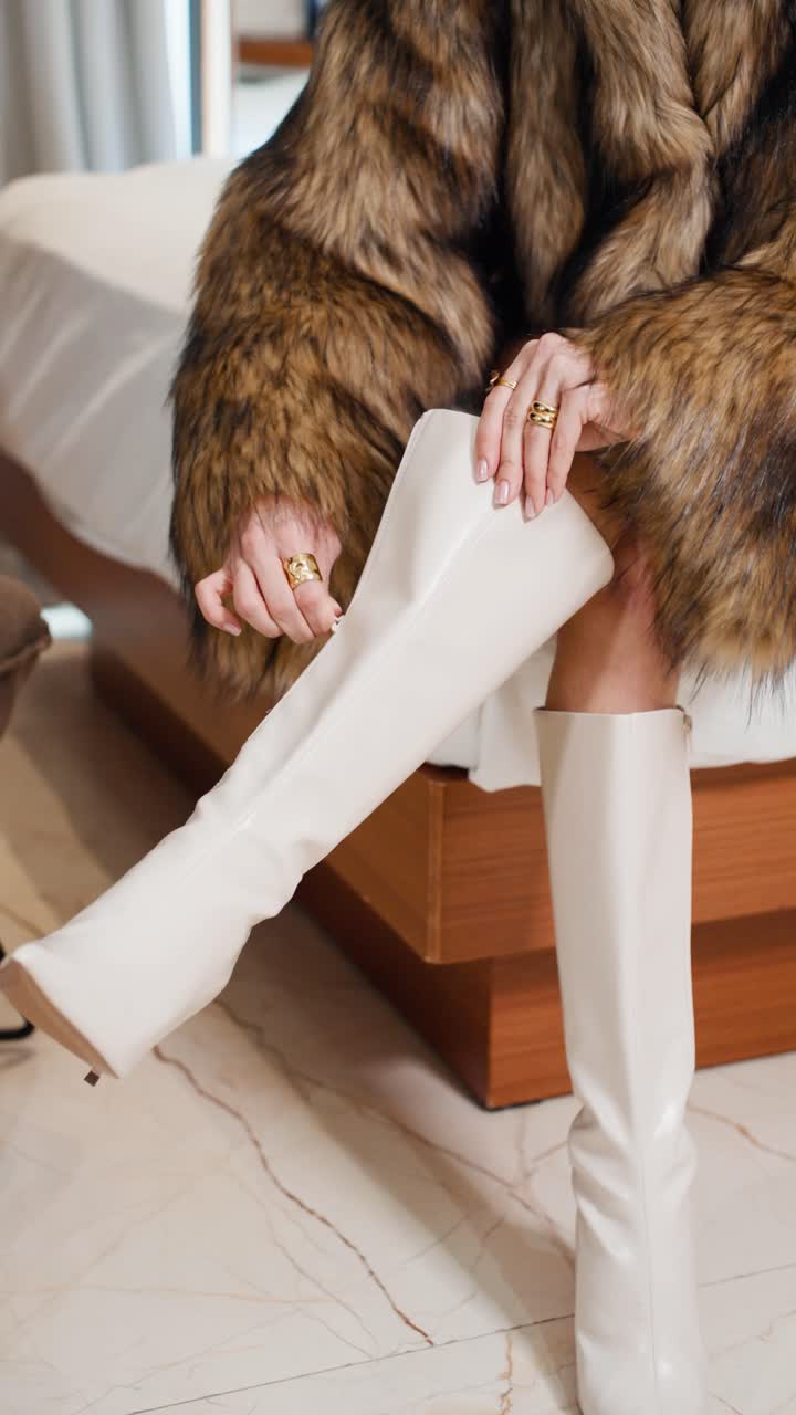 Fashinable woman wearing wearing a fur coat zip up knee-high cream boots while sitting on a modern wooden bed - close-up