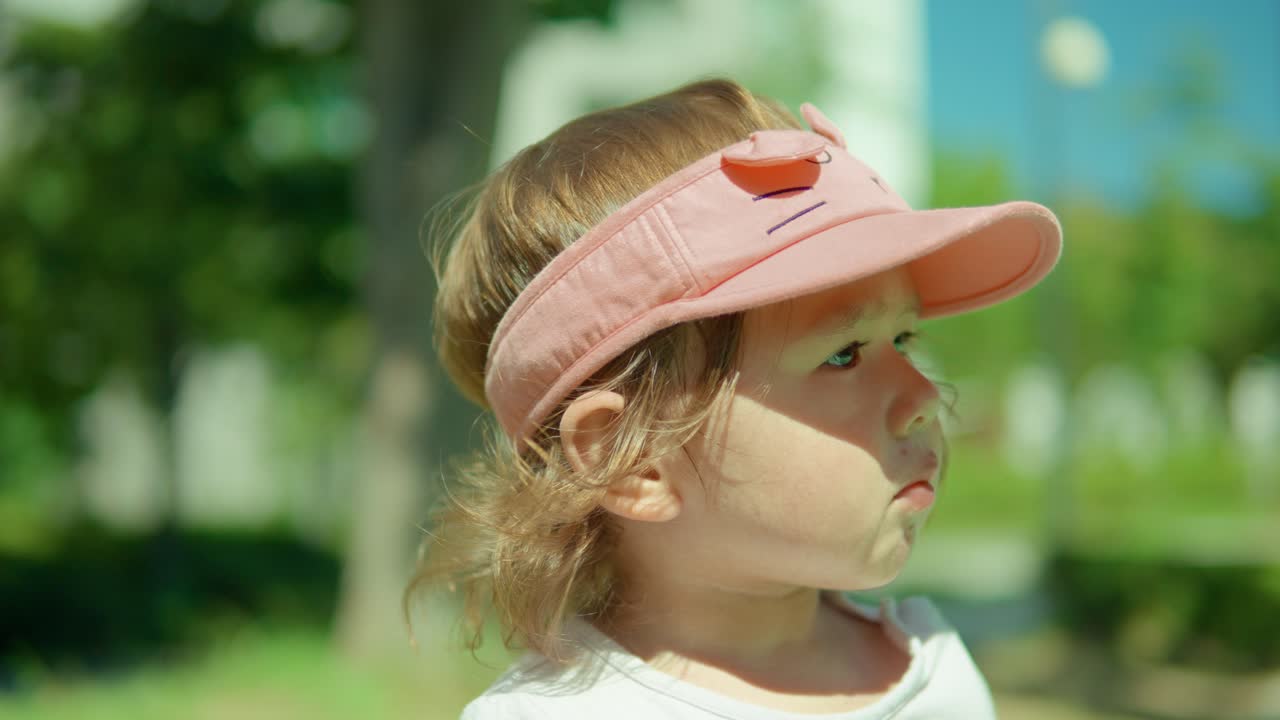 niña pequeña rubia con estilo masticando un sabroso refrigerio y usando un sombrero rosa mientras mira a un lado el parque al aire libre - retrato, cámara lenta