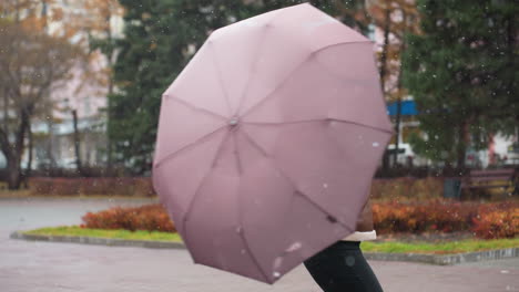 female holding umbrella, wearing knit cap, brown shearling jacket, black trousers, happily spinning umbrella in light snowfall, trees in background, enjoying autumn day, joyful and carefree