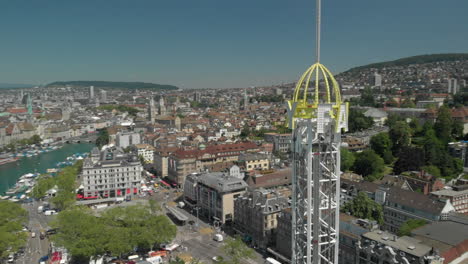 Disparo-Aéreo-De-Drones-Sobrevolando-Alrededor-De-La-Torre-De-Caída-Libre-Del-Parque-De-Diversiones-Con-La-Ciudad-Y-El-Lago-De-Zúrich,-Suiza-En-El-Fondo-Durante-El-Zürichfest