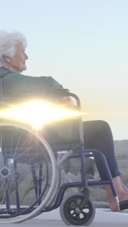 animation of light moving over smiling senior caucasian woman in wheelchair on beach looking to sea