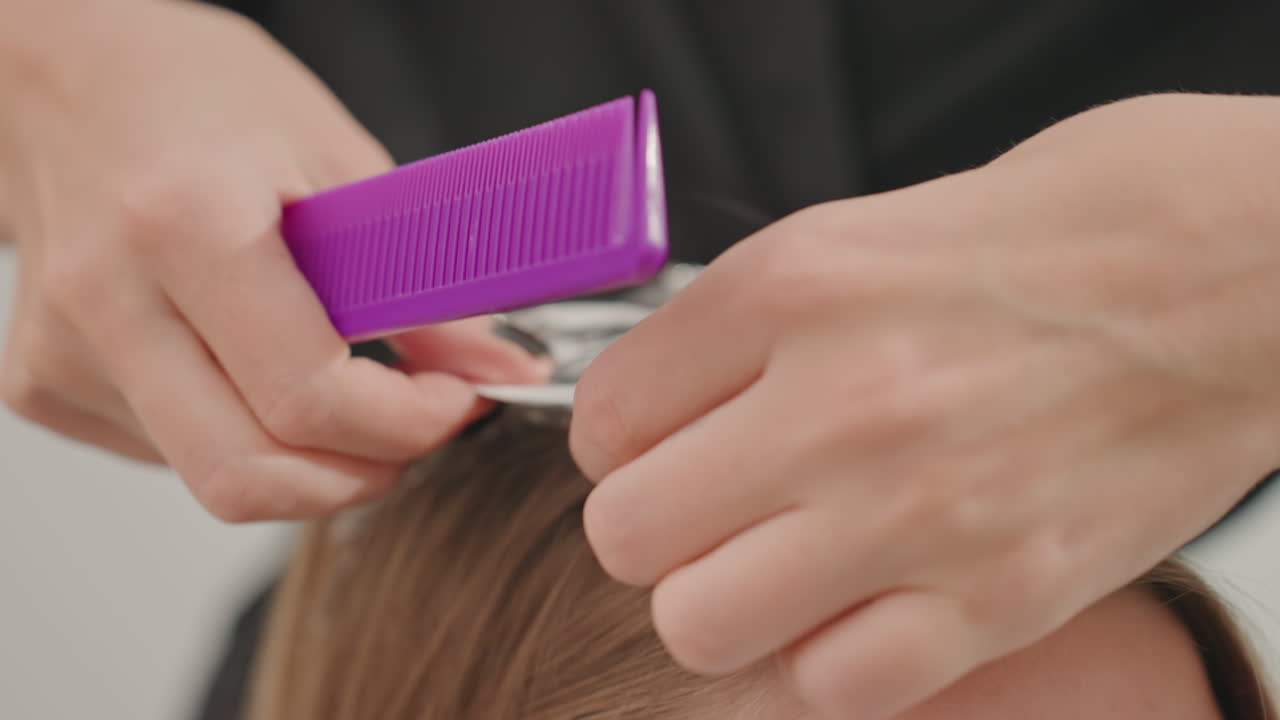 caucasian woman foiled highlights in salon, colorist carefully sections hair and applies dye to roots using purple comb and aluminum foil, closeup on hands with black nail polish, focused technique