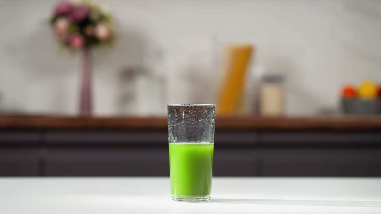 vaso medio lleno de jugo de pasto de trigo verde vibrante en un mostrador de cocina blanco, fondo borroso