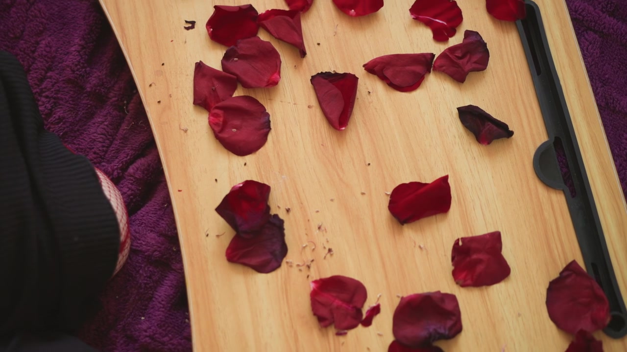 Rose petals on wooden board near seated woman performing fortune reading with candle and purple cloth, bohemian cozy ritual, closeup of scattered crimson petals on velvet, slow living aesthetic