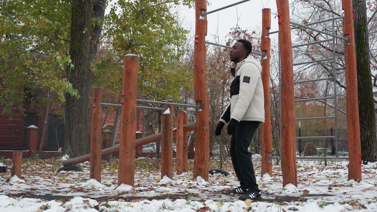 남자 는 겨울 에 나무 의 운동 장비 근처 에서 야외 에서 팔 의 유연성 운동 을 하며, 나무 와 금속 막대기 와 배경 에 주차 된 자동차 로 둘러싸인 눈 인 공원 에서 스트레칭 을 한다