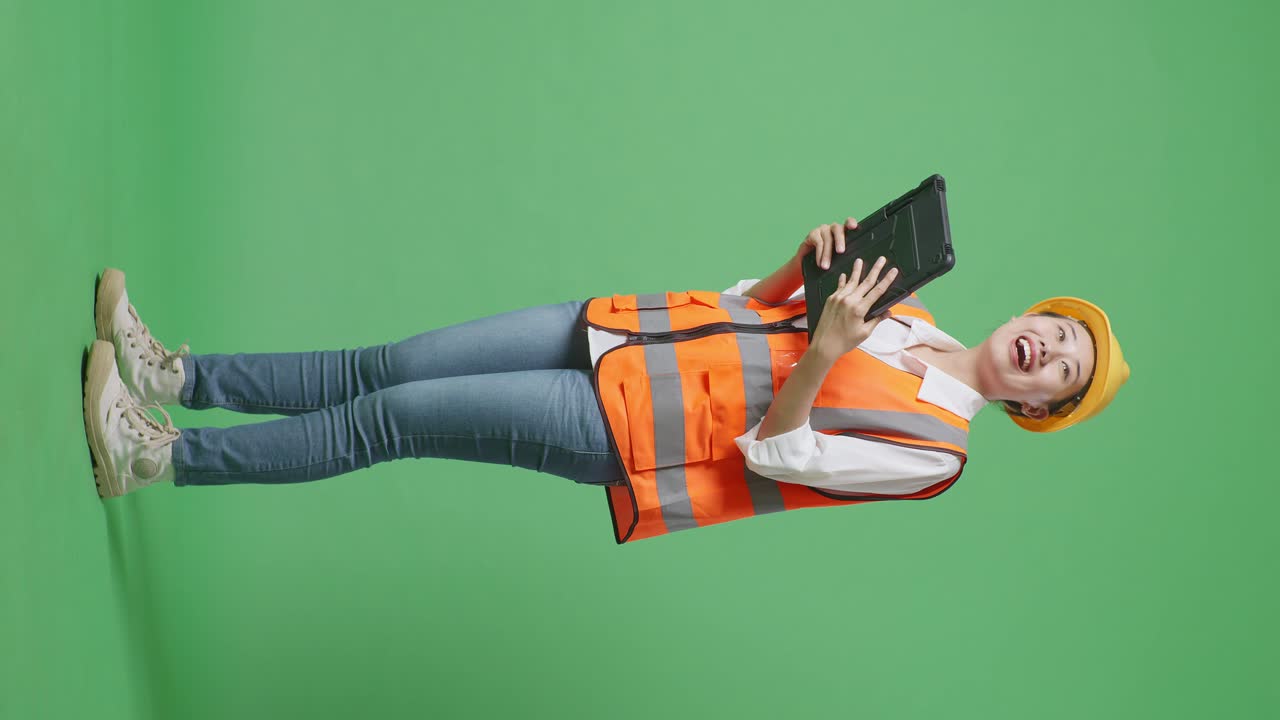 Full Body Side View Of Asian Female Engineer With Safety Helmet Looking At The Tablet In Her Hand And Looking Around While Standing In The Green Screen Background Studio
