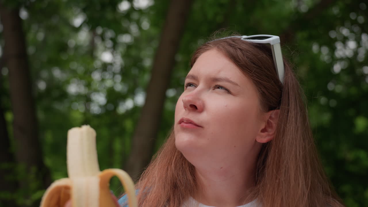 따뜻한 여름날, 젊은 여성이 나무 아래 공원 벤치에 앉아 자연광을 받으며 편안한 표정과 캐주얼한 복장으로 야외에서 신선한 바나나를 즐기고 있어요.