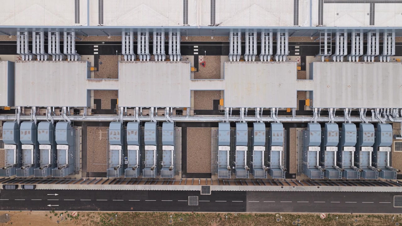 Rækker af kølesystemer er i drift på taget af et stort industrielt datacenterkompleks på en klar dag med asfalterede veje synlige omkring infrastrukturbygningens omkreds