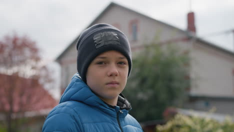 close-up of boy wearing black beanie and blue jacket standing outdoors, closing and opening eyes thoughtfully, with blurred red-roofed house, autumn foliage, and serene outdoor surroundings