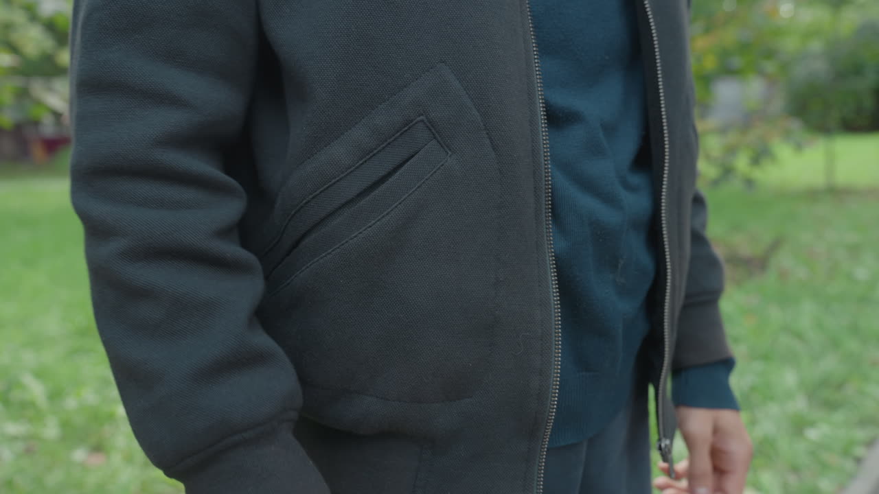 Uomo bianco ripreso a mezzo busto che sistema la tasca della giacca, atteggiamento rilassato da pendolare su un sentiero del parco, tasca in maglia lavorata, mani che scivolano nella tasca, atmosfera mattutina rilassata con prato verde sullo sfondo e spontaneo