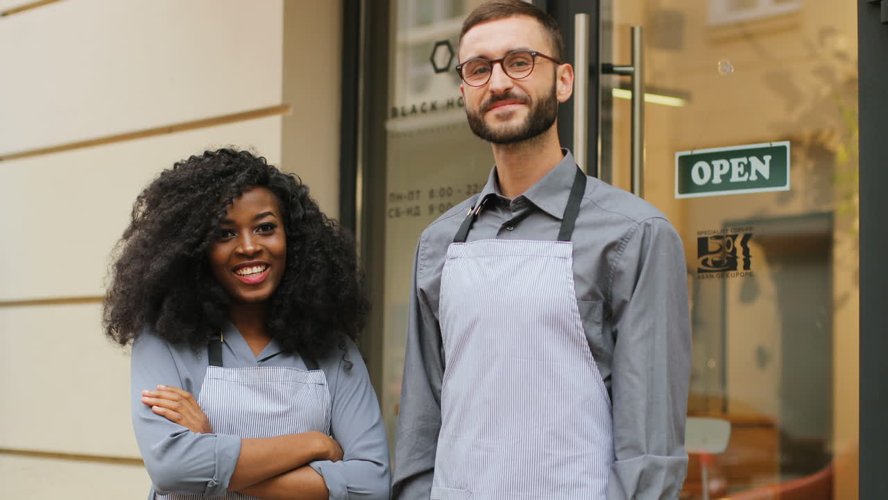 barista caucásico y barista afroamericana de pie en la puerta de un café moderno, sonriendo y mirando la cámara