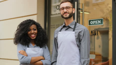 caucasian male barista and african american female barista standing on the door of a modern cafe, smiling and looking at the camera