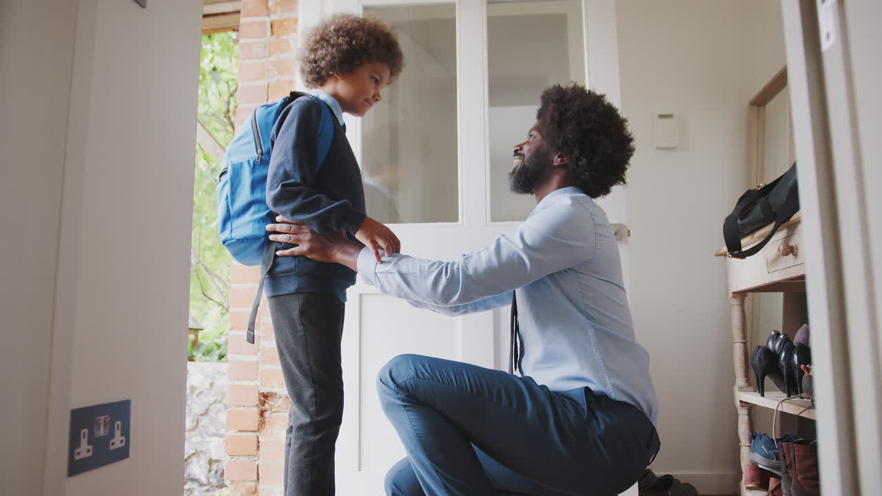 Middle aged black man wearing a shirt and tie kneeling down preparing his son and saying goodbye before he leaves home for school in the morning, side view