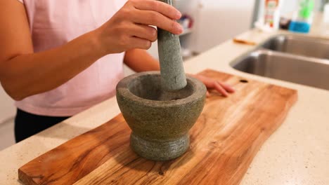 a person grinds ingredients with a mortar and pestle on a wooden board in a well-lit kitchen