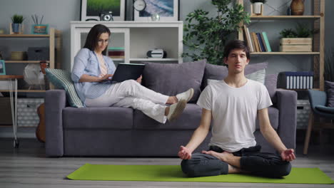 couple relaxing at home, man meditating, woman working on laptop.