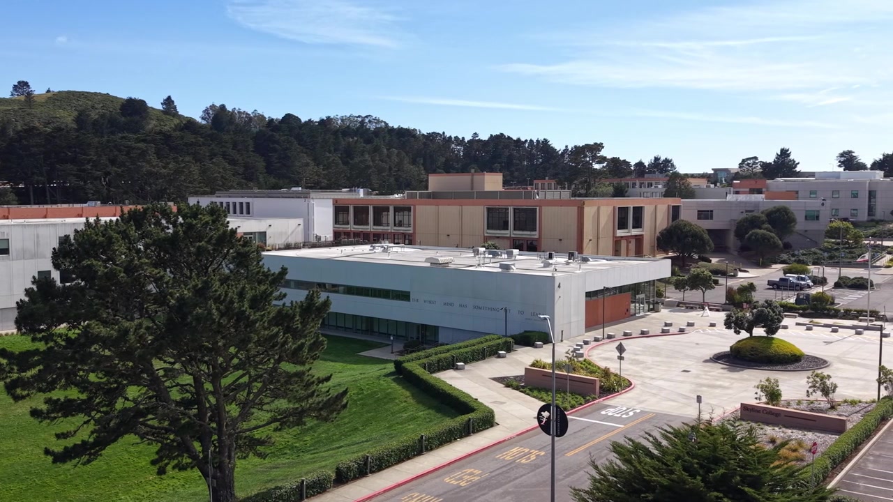 Dron aéreo sobrevolando el campus de Skyline College en San Bruno, California, mostrando arquitectura moderna, zonas verdes, áreas de estacionamiento y colinas circundantes bajo un cielo azul claro.