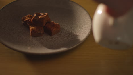 close up of person pouring out steaming cup of hot chocolate drink on table next to plate of brownies