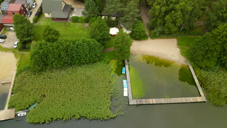 Sandy-beach-of-Hartowiec-Lake-with-boats-floating-near-the-wooden-jetty-on-a-sunny-day-in-Poland,-Aerial-take-off-top-down-view,-small-private-hotels-and-houses-on-background