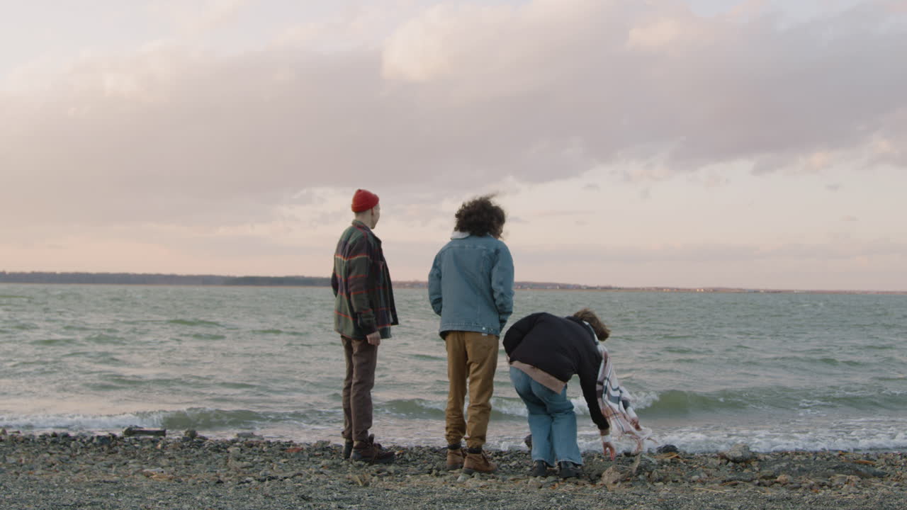 겨울 옷을 입은 세 친구가 흐린 날 바닷가에서 자갈을 물에 던지고 이야기하는 뒷모습