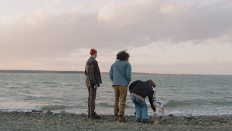 rear view of three friends in winter clothes talking and throwing pebbles to the water on a seashore on a cloudy day