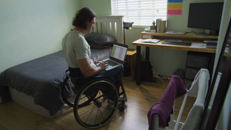 hombre con discapacidad caucásico enfocado en silla de ruedas usando computadora portátil en el dormitorio