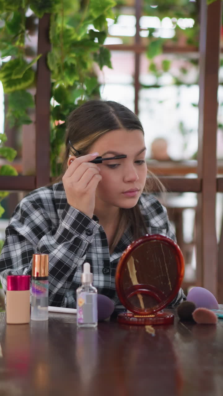 Mesa con espejo, rímel y arreglo de cejas entre vegetación. Una adolescente se aplica rímel y se arregla las cejas en la mesa de una cafetería adornada con plantas, con un espejo de bolsillo y frascos colocados sobre madera, en un delicado ritual matutino de cuidado personal.