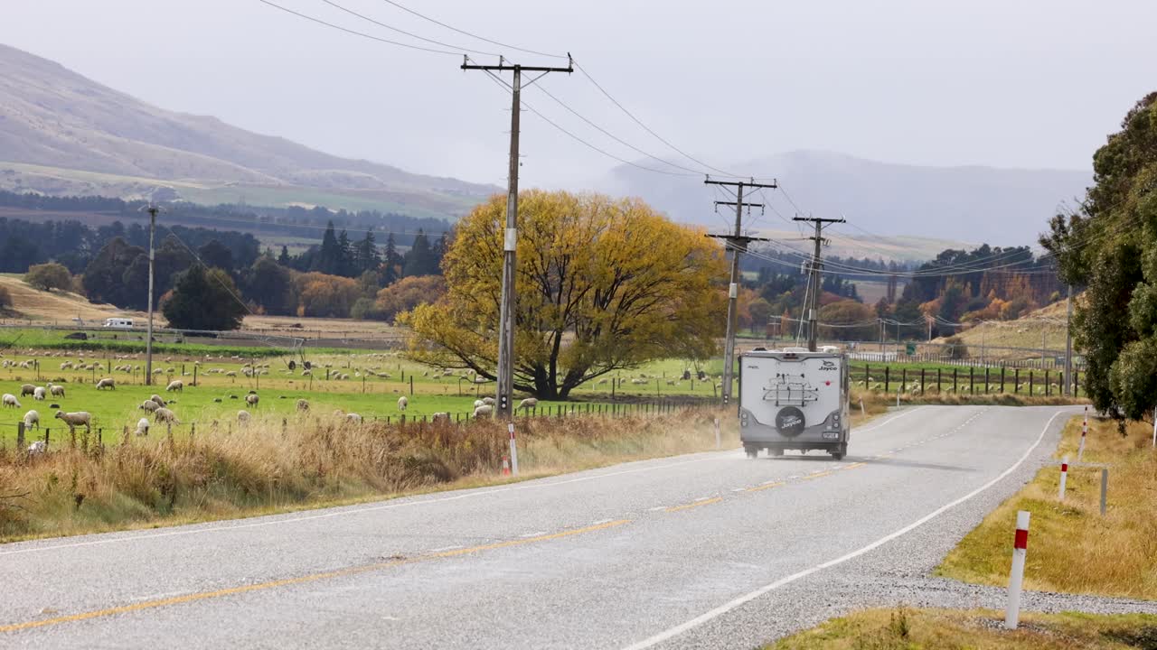 A vehicle travels along a rural road in Wanaka, New Zealand, surrounded by lush greenery and distant mountains under soft daylight