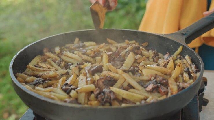 Camping skillet sizzling potatoes and mushrooms outdoors, hands stirring with wooden spatula over portable gas stove, steam rising against grassy lakeside background, rustic camp cook preparing crispy