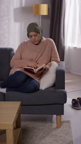 Vertikales-Video-Einer-Muslimischen-Frau-Mit-Hijab,-Die-Zu-Hause-Auf-Dem-Sofa-Sitzt-Und-Den-Koran-Liest-Oder-Studiert