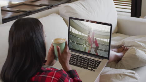 compuesto de una mujer sentada en casa sosteniendo un café viendo un partido de rugby en una computadora portátil